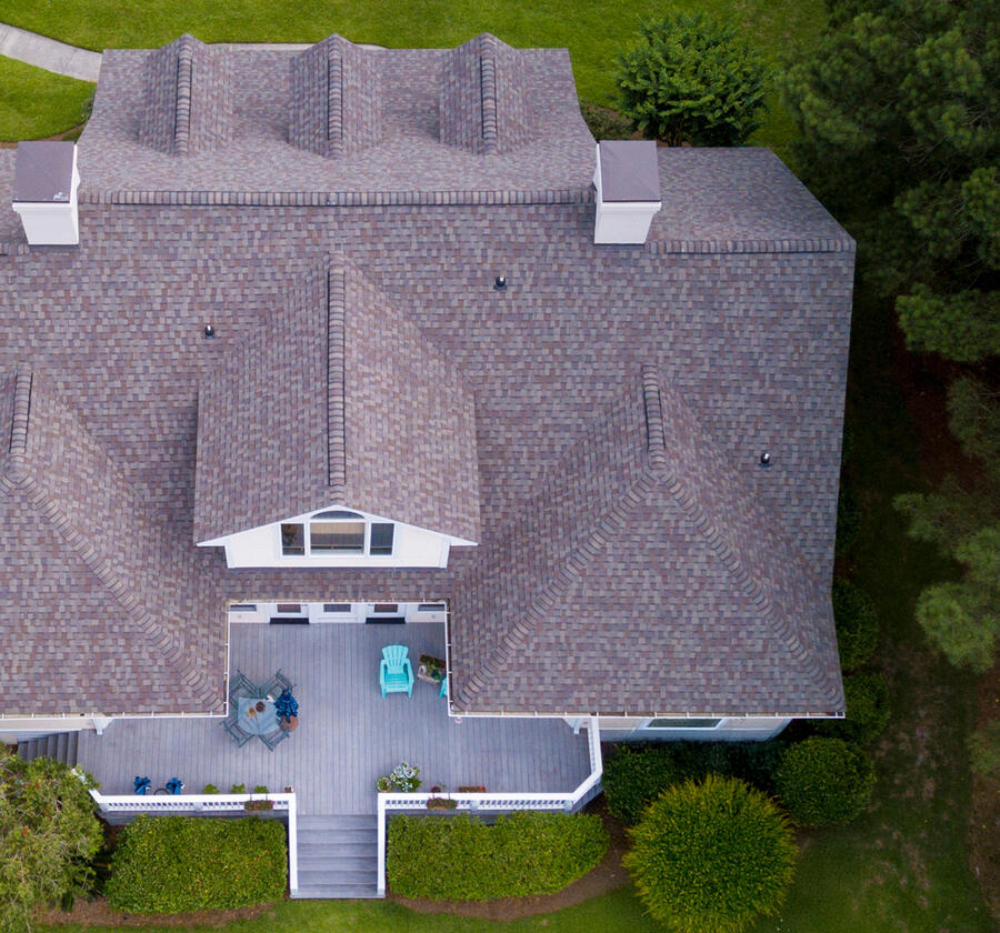 Aerial view of a house with a shingled roof, a backyard patio with outdoor furniture, and surrounded by trees and green lawn.